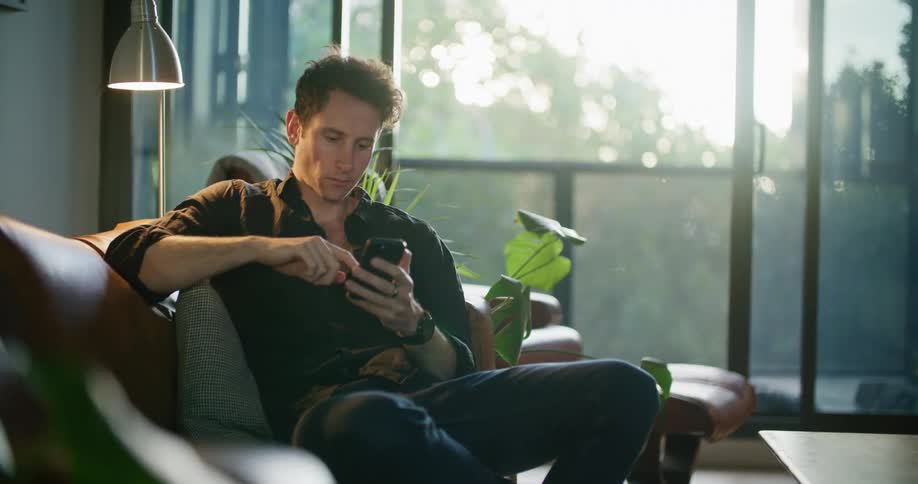 Man with black shirt and jeans sits on brown couch using smartphone in living room at home