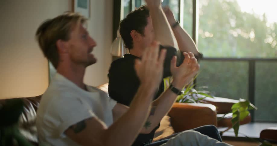 A pair of men cheer and clap while watching television together in the living room of a house during the day