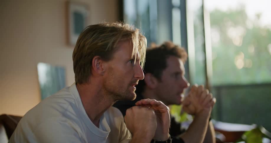 Two men are sitting on a couch watching television they cheer at the same time