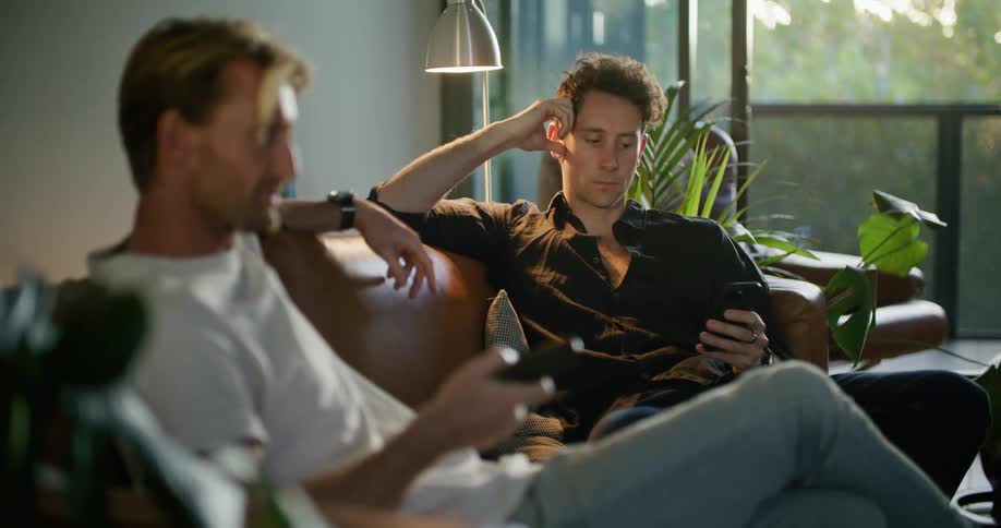 A man uses a remote control for a television while his friend sits on the same couch scrolling through his phone