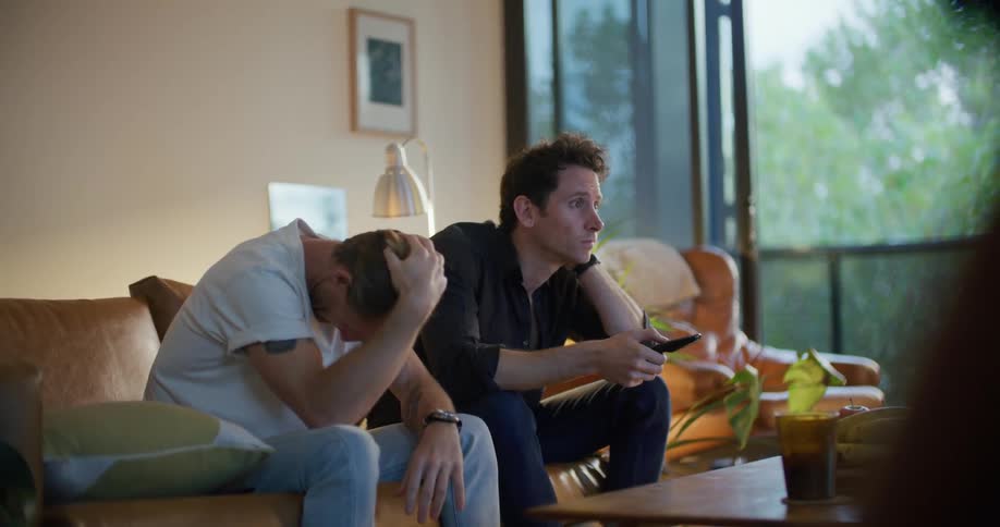 Two men sit on a couch and talk and point as they watch television