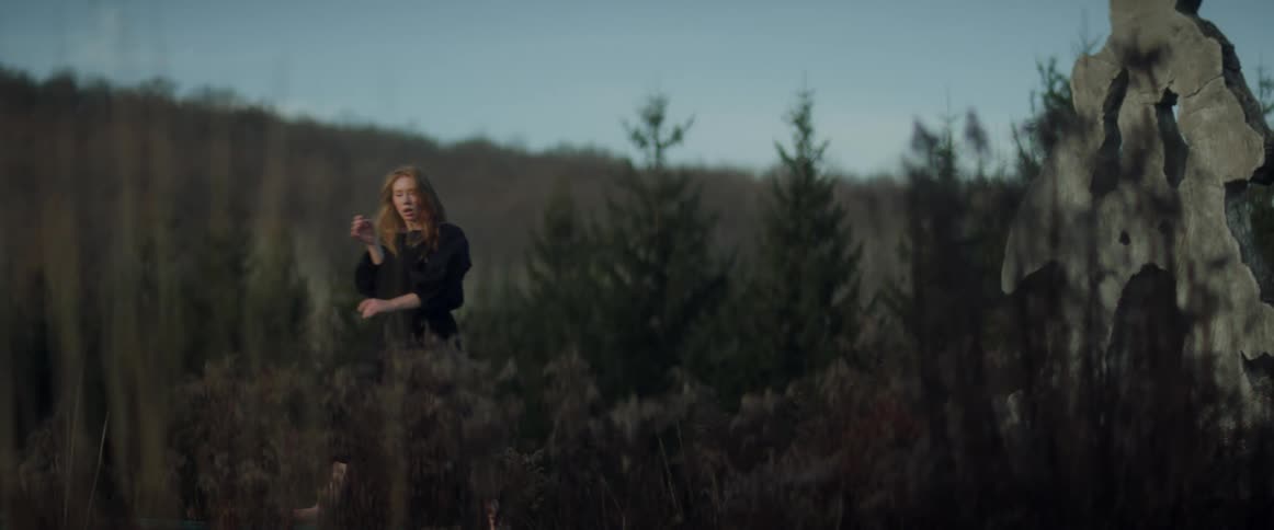 A teenage girl wearing a black dress is outside dancing near trees and a face statue with forests in the distance