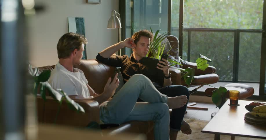 Two men wearing jeans white and black shirts sits on brown couch using smartphones in living room at home