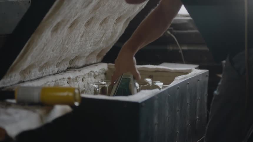 A glass blower uses tongs to place a piece of glass into a cooler