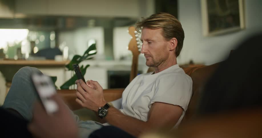 A man sitting on a sofa scrolling and tapping on his phone turns his head to talk to a friend doing the same