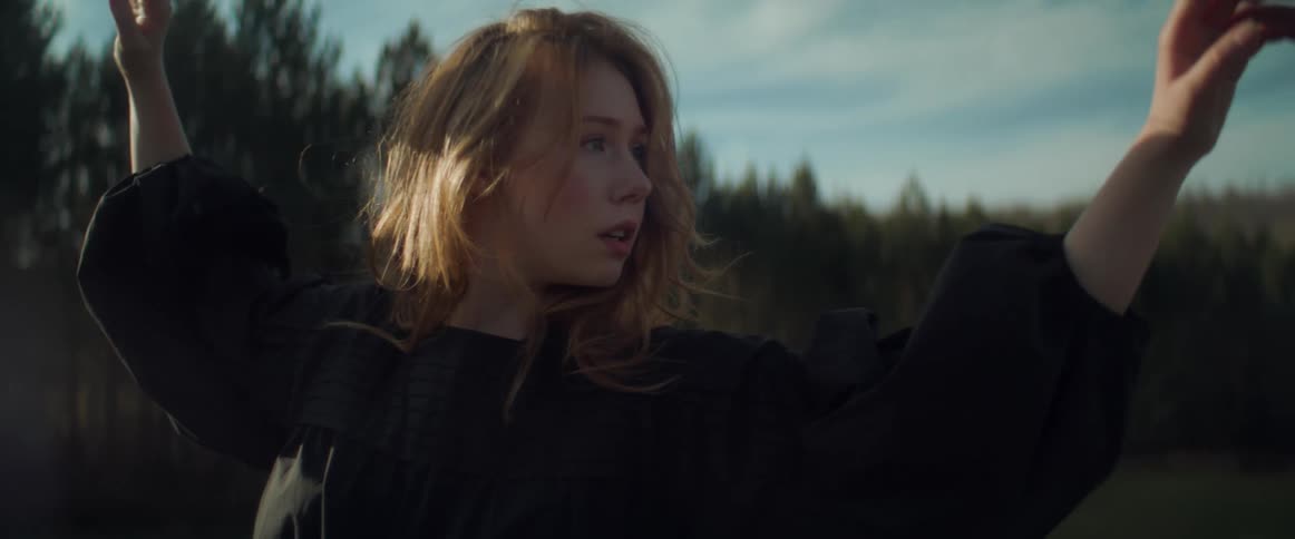 A teenage girl with long hair and wearing black is outside dancing and there is a forest of trees in the distance