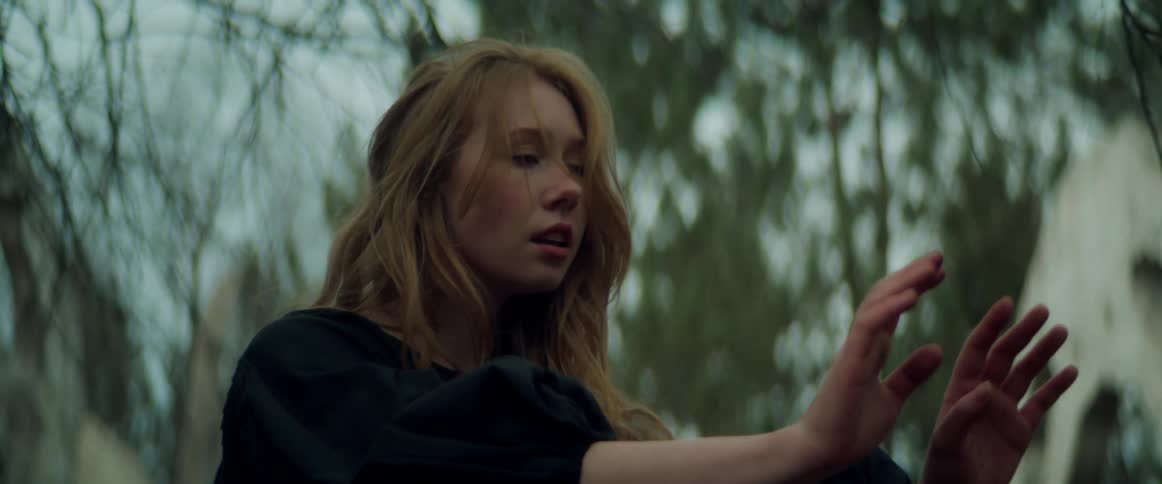 Blonde woman with wavy hair and black dress dances in the forest moving her hands