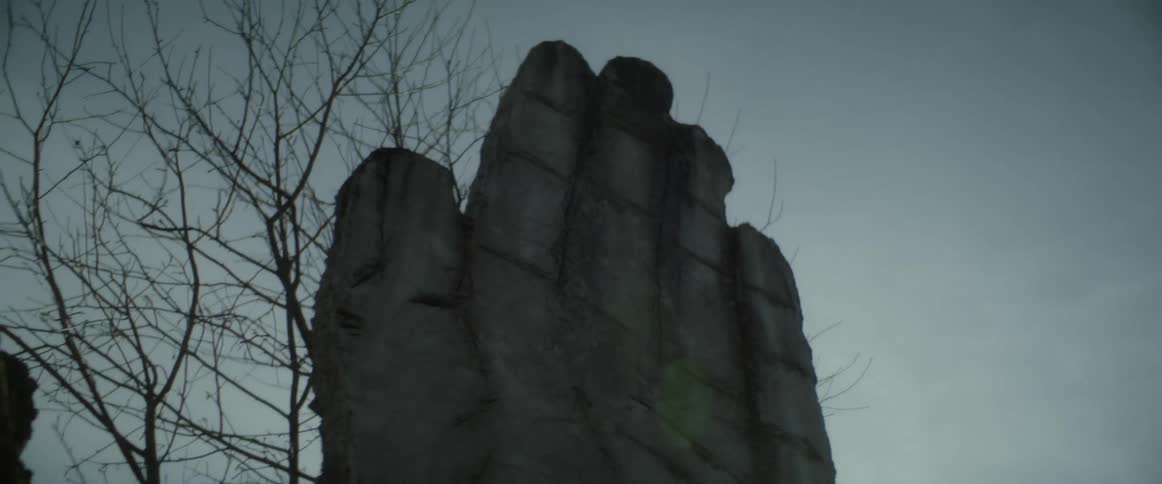 The sun shines behind large carvings of a hand and a face near a bare tree and there is a forest in the distance
