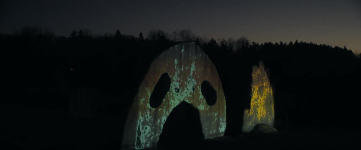 Flat metal sculptors lit by projected images stand in row backed by forest covered hills at night