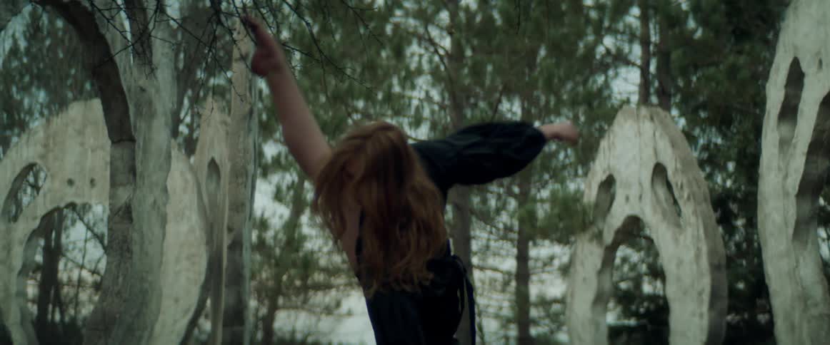 A girl wearing a black dress is outside dancing in front of stone face carvings surrounded by trees and a forest in the distance