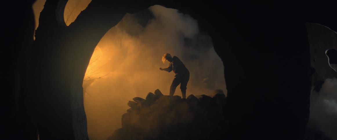 Woman wearing black clothes dances over a mound of rocks surrounded by sculptures and smoke at night