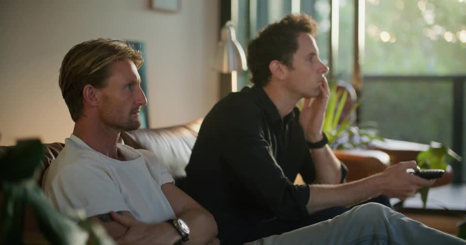 Two men are sitting on a couch and one man holds up a remote controller and pushes a button