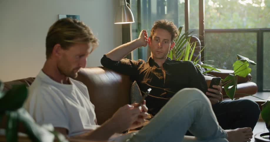 Two men wearing jeans white and black shirts sits on brown couch using smartphones in living room at home