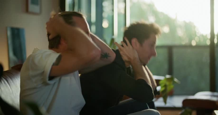 Two men are sitting on a couch watching an unseen television and both men put their hands on their faces and appear frustrated