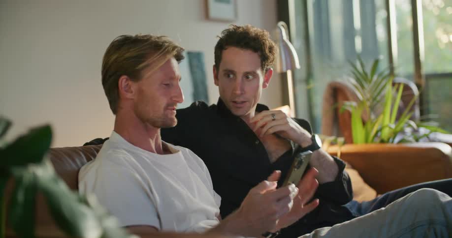 Two men are sitting on a living room couch talking as one man is showing the other man something on his phone