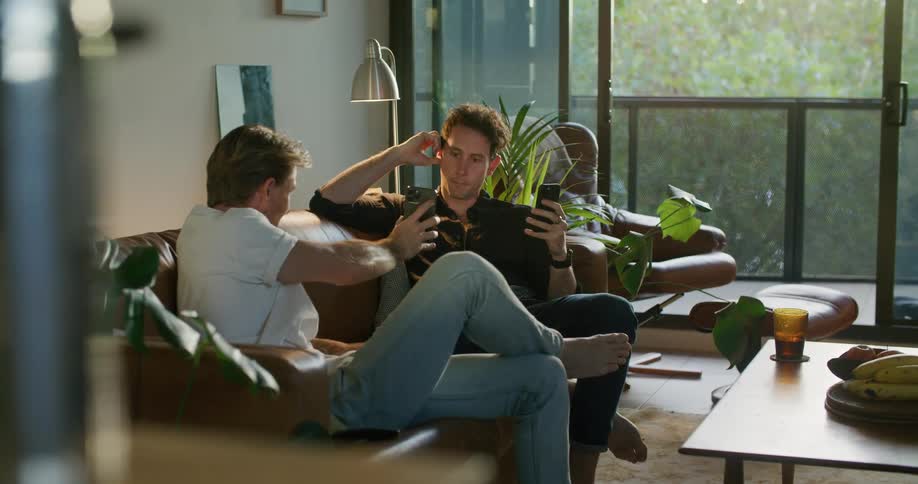 Two men sit on a couch in a home as they both look at their cell phones and then on mans leans over and shows his phone to the other