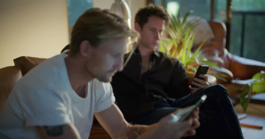 Two friends are sitting on a couch and looking at their cell phones