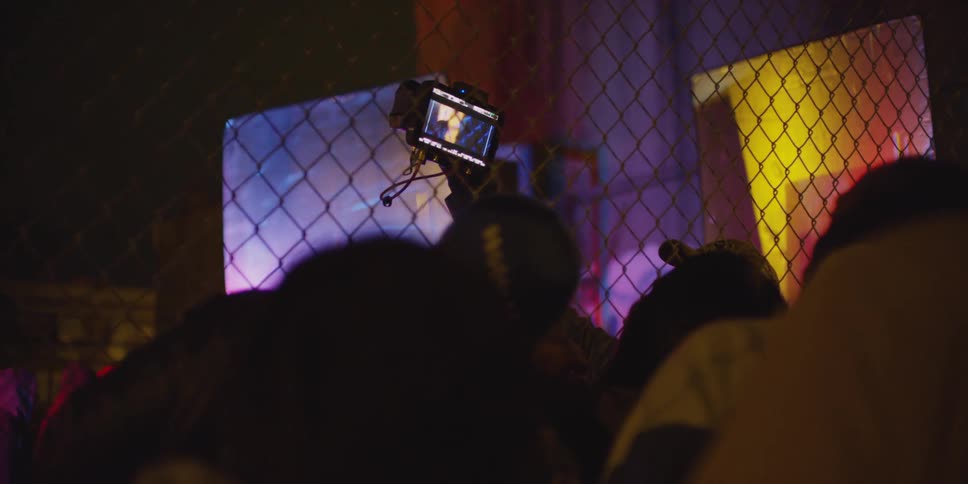 A crowd stands next to a chain link fence at night while one person holds a video recorder in the air