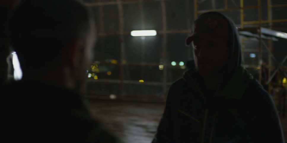 A film crew member walks up to someone in a court at night