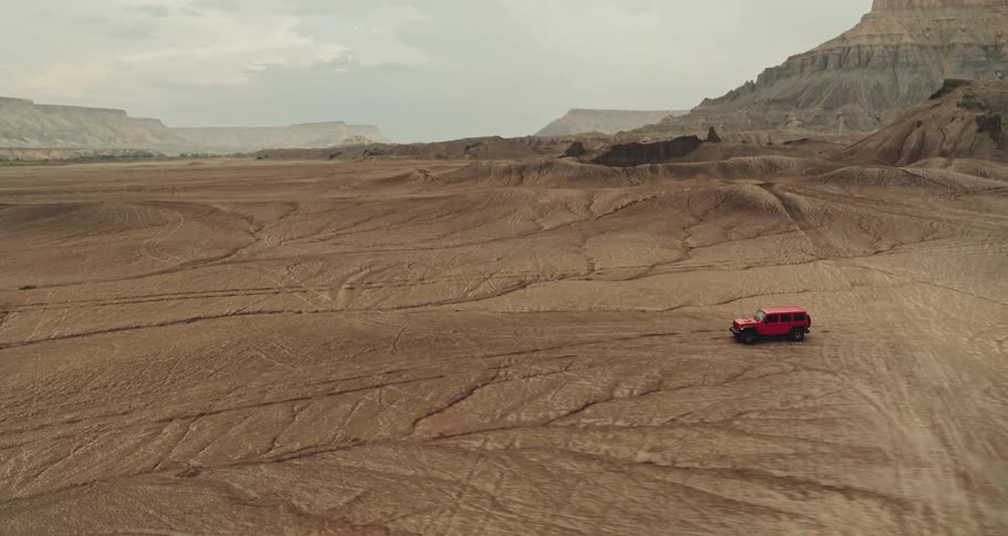 A red truck travels across the desert with mountains and mesa in the distance under a cloudy sky