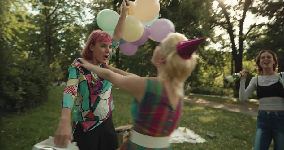 Two teenage girls carrying balloons champagne and gifts walk to a park and hug another girl wearing a party hat
