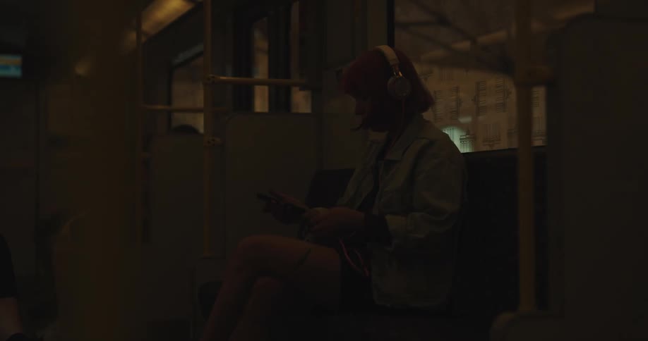 A teenage girl with pink hair is sitting in a seat on a train and she puts her headphones on her head
