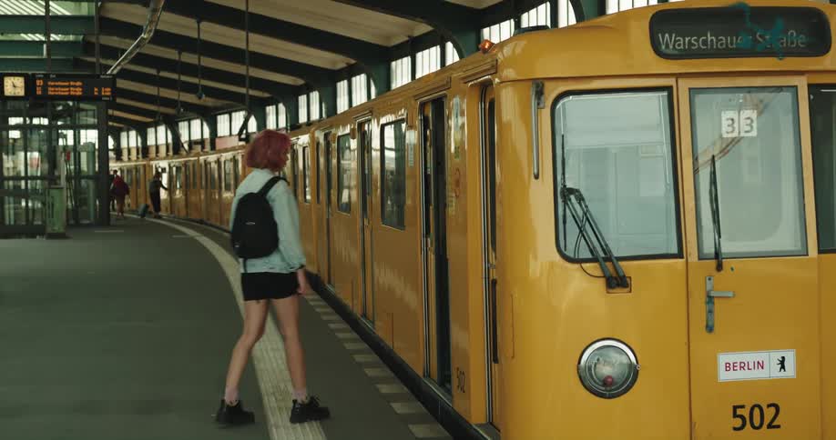 A teenage girl with a backpack on her shoulder is standing at a train station and she walks on to a train after passengers get off it