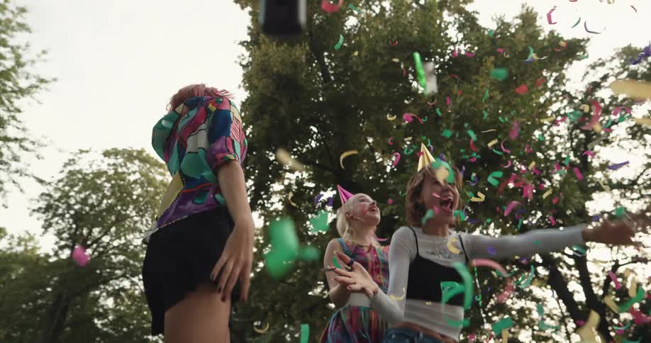 Female friends jump and dance outdoors spreading confetti and wearing party hats