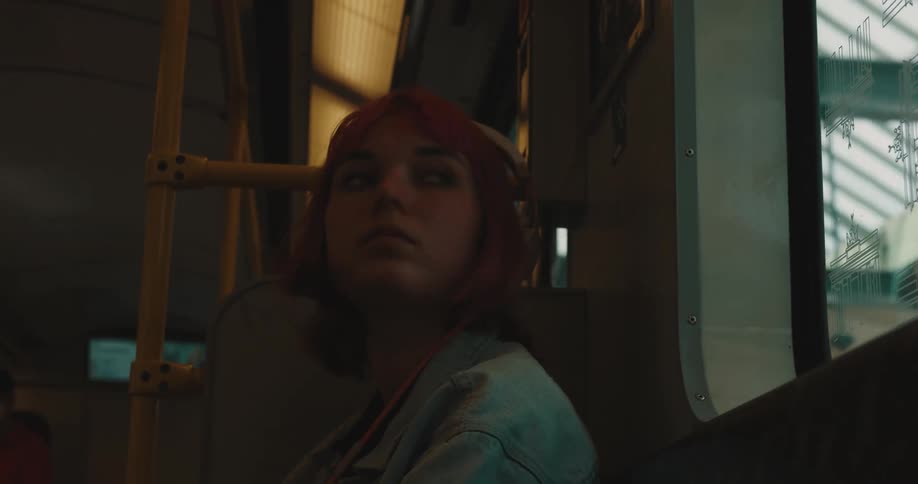 A teenage girl with pink hair and wearing headphones is sitting on a train and she gets up and walks out when it stops