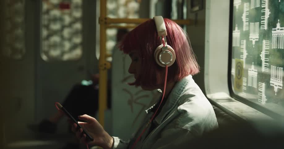 A woman with pink hair who is riding a bus puts on headphones and is listening to music