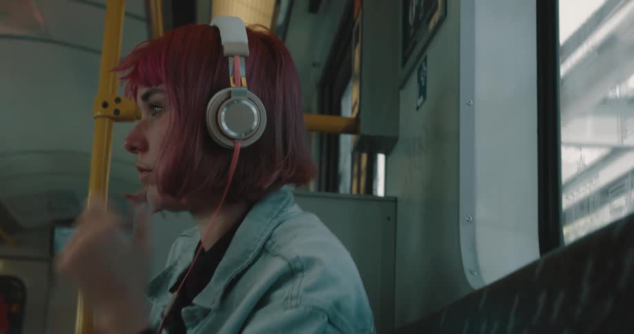 A teenage girl with pink hair is sitting in a seat on a train and she puts her headphones on her head