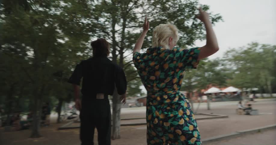 A woman and man dance together atop a bench in a public park during the day