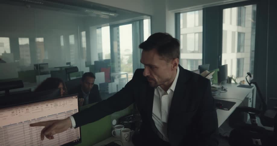 Man leans against desk discussing data on screen with woman in office