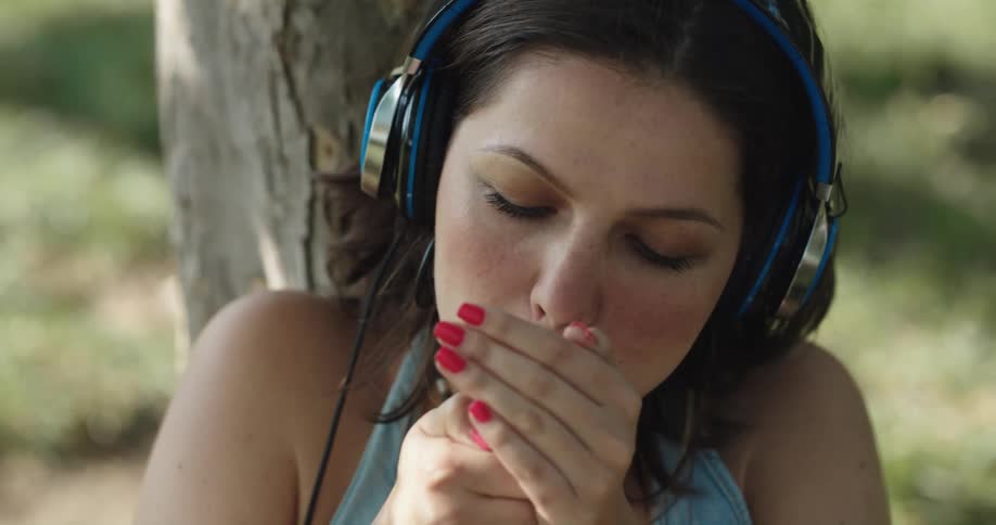 A woman sitting outdoors against a tree while wearing headphones places a cigarette in her mouth and lights it with a lighter