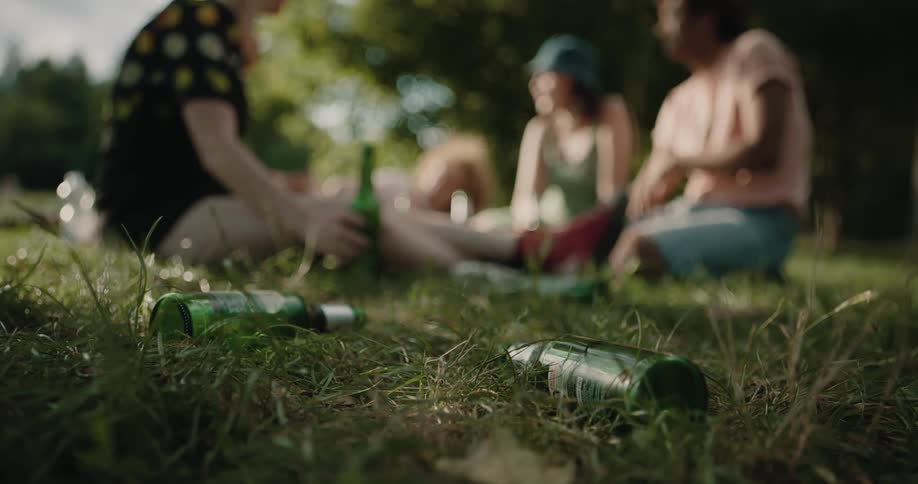 A woman tosses an empty beer bottle on the ground while picnicking with friends in the park