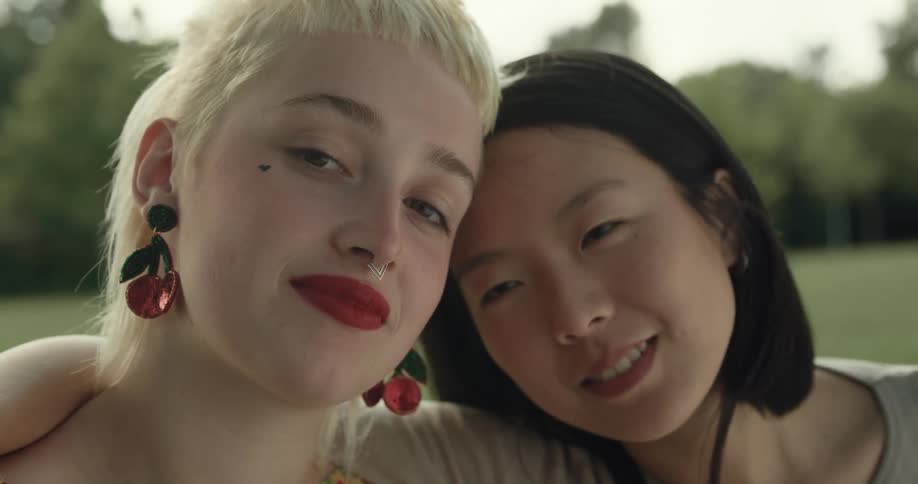 A pair of women sitting outdoors with their arms around one anothers shoulders look into the camera for a moment before turning to smile at one another