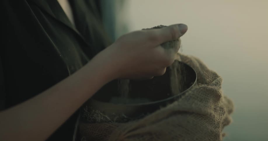 A woman reaches in to a container and picks up a handful of ashes and holds them in her hand and then tosses them in to the air