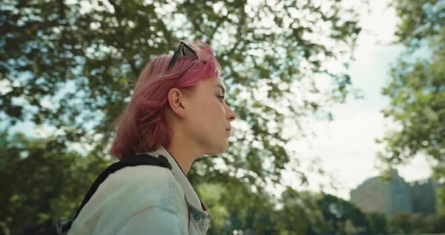 A teenage girl rides a scooter down a path in a public park past a woman and her dog