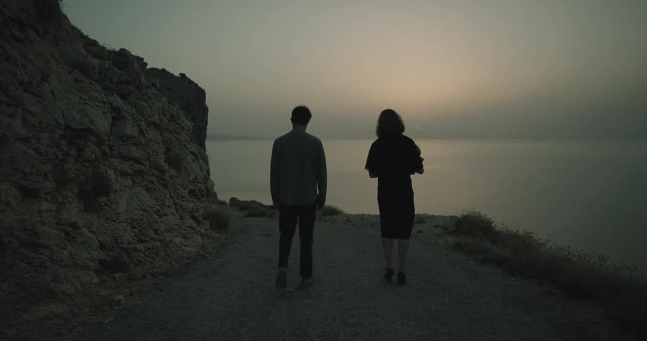 Silhouettes of two people walking on a mountain road along the coast as the sun sets over the water