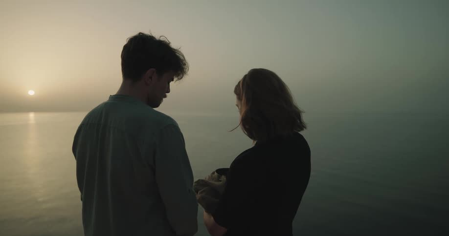 A woman opens an urn and hands the cover to a man standing next to her and scatters ashes into the sea