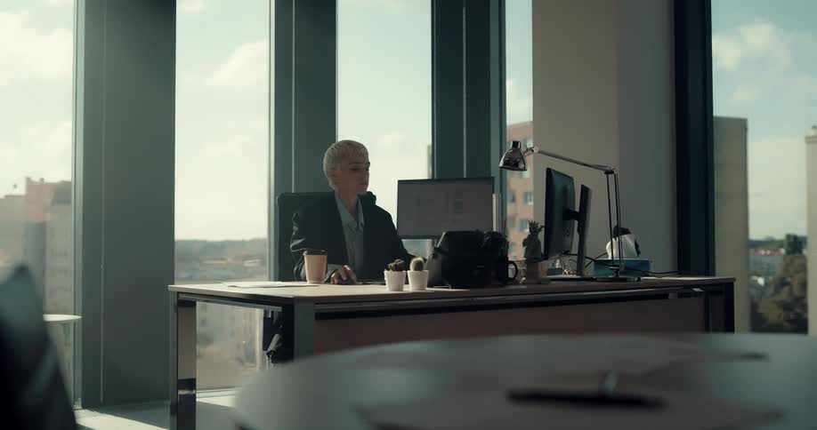 A woman walks into her office drinking coffee and sits down at her desk