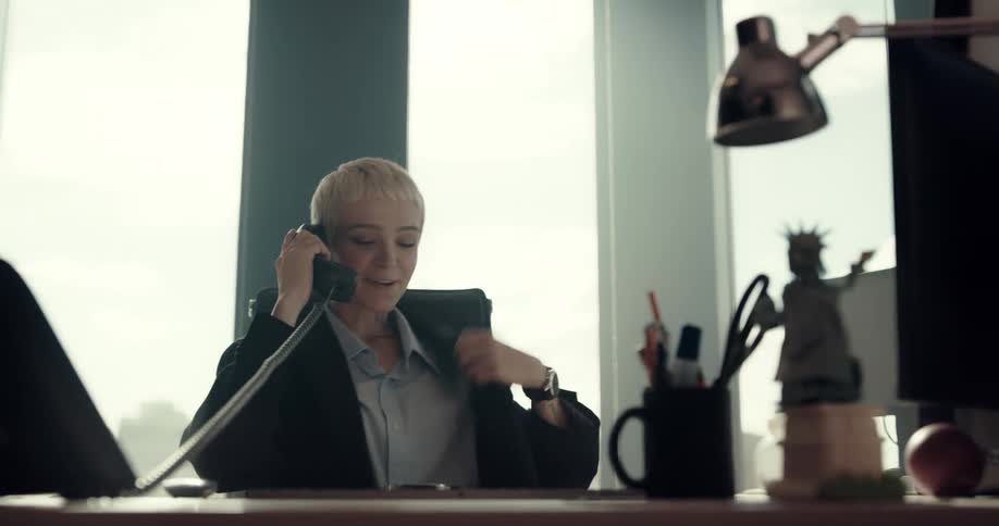 A woman laughs and looks at her watch as she sits at a desk on the phone