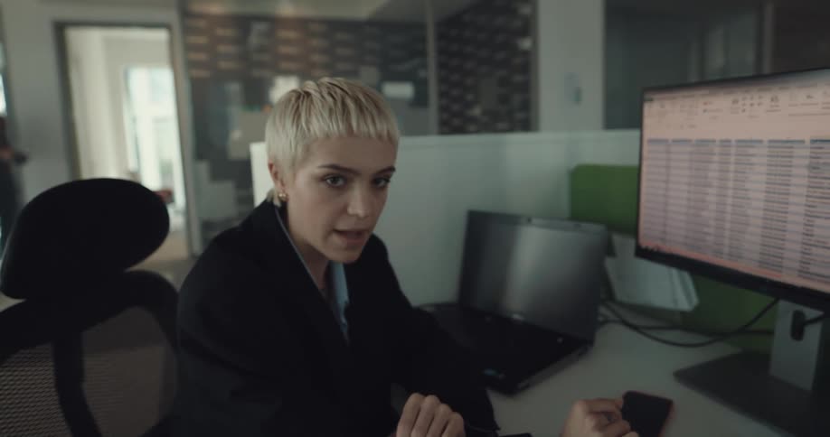 A woman is sitting at a desk in front of a computer talking and pointing at the monitor