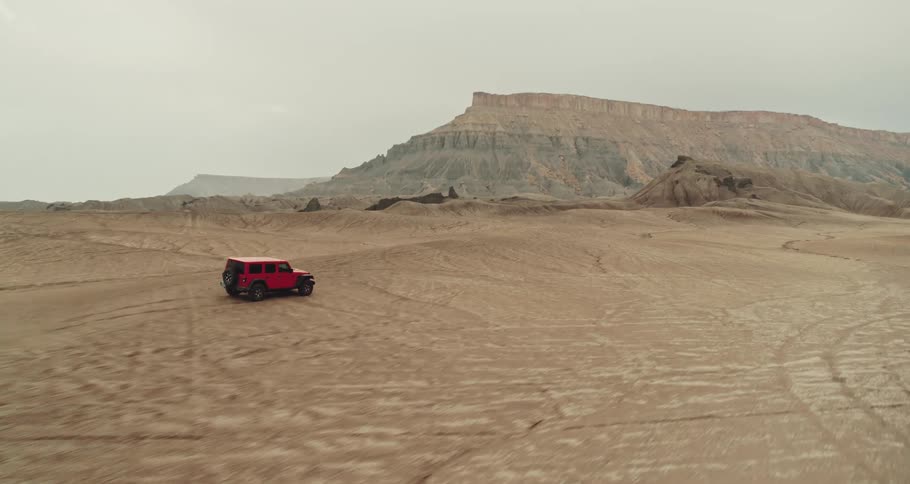 A red truck travels across the desert with mountains and mesa in the distance