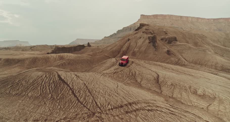 A red truck travels on rough terrain in the desert badlands among mountains and mesa under a cloudy sky