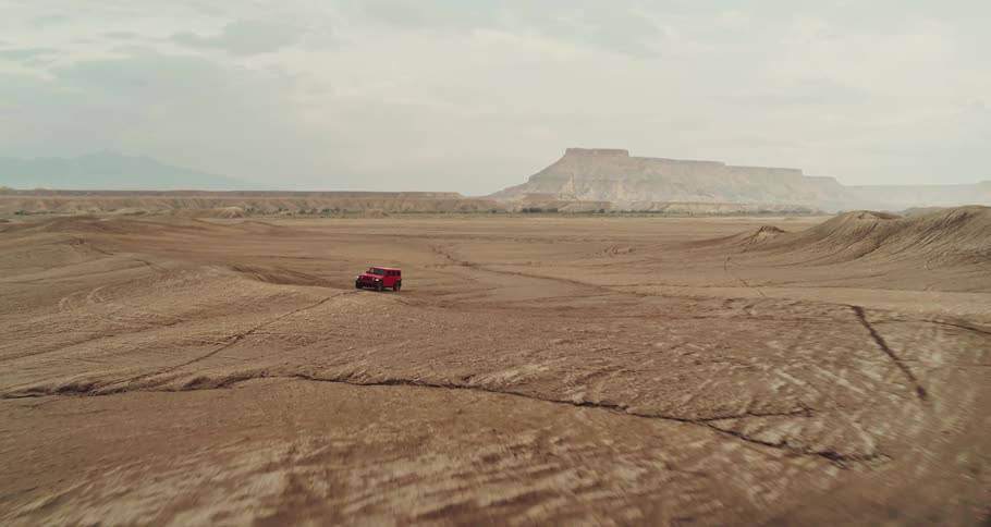 Red jeep rides over uneven desert terrain stretching toward mist covered mountains