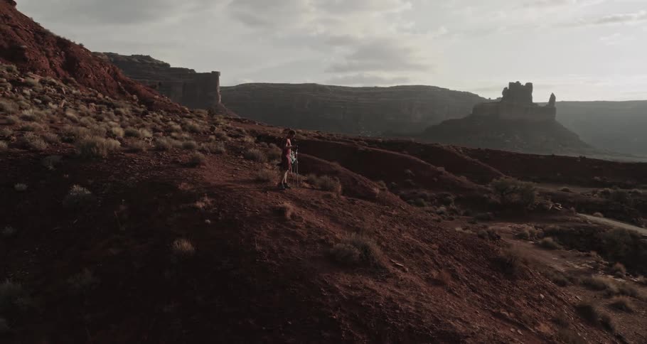 A man is standing on a hill overlooking the desert badlands as the sun shines brightly upon buttes and mountains