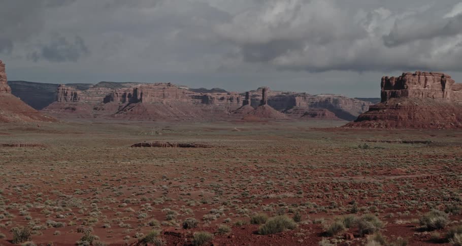A hiker is standing in the desert badlands holding trekking poles and there are buttes and mesa in the distance under a cloudy sky