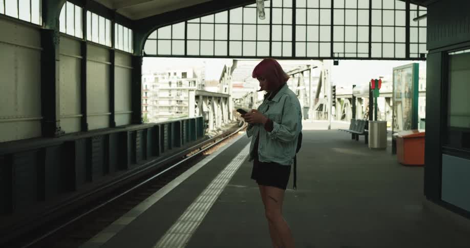 A teenage girl with a backpack on her shoulder is standing at a subway station looking down the tracks for her train and using her cell phone