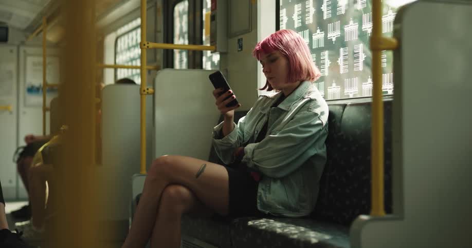 A teenage girl with pink hair is sitting on a train texting on her phone and looking out the window